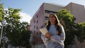 Eine Studierende geht lächelnd mit Einschreibungsunterlagen im Arm vor der Universität.